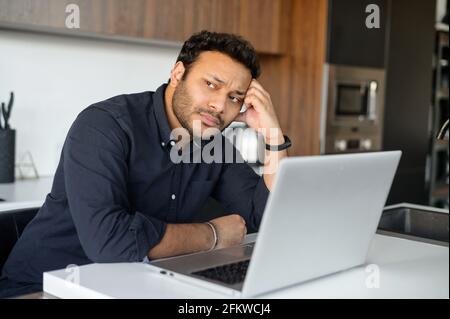 L'uomo disoccupato indù depresso sta usando il laptop per la ricerca di lavoro, il giovane ragazzo indiano penoso sconvolto si siede in cucina a casa con il laptop, si riposa con la testa con la mano, si guarda via, si sente triste e preoccupante Foto Stock