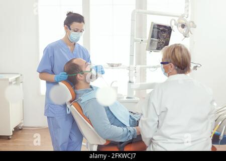 Assistente medico che mette la maschera di oxigen che esamina il paziente malato dell'uomo per la chirurgia di odontoiatria mentre sta in piedi sulla sedia dentale nella stanza della clinica di stomatologia. Dentista medico che esamina toohache Foto Stock