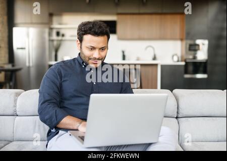 Sorridente allegro ragazzo freelancer misto-corsa sta usando il computer portatile seduto sul divano a casa, ragazzo indù gode di lavoro remoto da casa in un'atmosfera confortevole. Il maschio indiano spende il tempo libero in linea Foto Stock