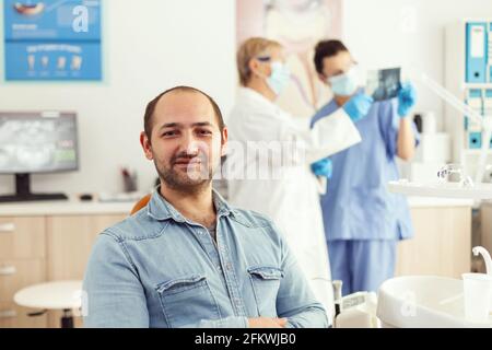 Paziente uomo che guarda in macchina fotografica in attesa di risultati radiografici seduto su sedia dentale in ospedale stomatologia clinica. In background dentisti squadra che guarda i denti x-ray preparazione per l'intervento chirurgico Foto Stock