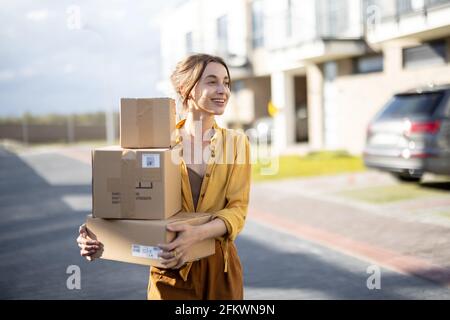 Giovane donna felice porta a casa un pacchi con merci acquistate online nel moderno quartiere residenziale. Concetto di acquisto e consegna online Foto Stock