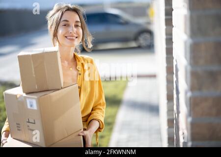 Giovane donna felice porta a casa un pacchi con merci acquistate online, bussando alla porta. Concetto di acquisto e consegna online. Guardando la fotocamera. Foto Stock