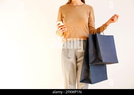 Giovane donna snella con un classico outfit alla moda, un pullover marrone e pantaloni grigi sciolti sono andati a fare shopping, tenere borse di carta e chiacchierare sullo smartphone. Bianco Foto Stock