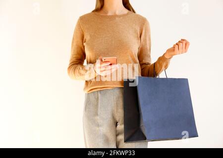 Giovane donna snella con un classico outfit alla moda, un pullover marrone e pantaloni grigi sciolti sono andati a fare shopping, tenere borse di carta e chiacchierare sullo smartphone. Bianco Foto Stock