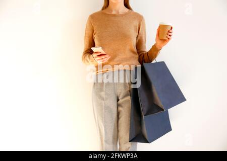 Giovane donna slim con abito classico alla moda, pullover marrone e pantaloni grigio sciolto è andato a fare shopping, tenere borse di carta, chiacchierare su smartphone, bere Foto Stock