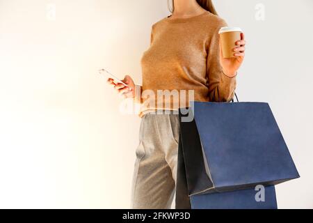 Giovane donna slim con abito classico alla moda, pullover marrone e pantaloni grigio sciolto è andato a fare shopping, tenere borse di carta, chiacchierare su smartphone, bere Foto Stock