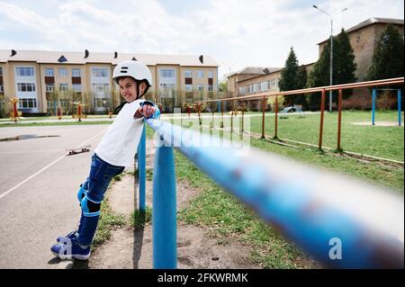 Bel ragazzo a tutta lunghezza con casco di sicurezza e attrezzatura protettiva appoggiata su una barra orizzontale sul parco giochi all'aperto. Concetto di attività per il tempo libero, sport, ad es Foto Stock