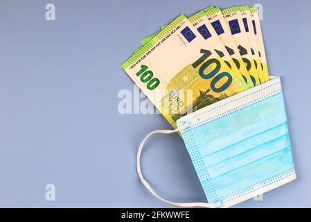 Maschera medica e denaro contante. Banconote in euro avvolte in maschera chirurgica monouso protettiva su sfondo grigio Foto Stock
