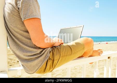 Calzare il giovane con il computer portatile seduto su veranda in legno bianco. Vantaggi del concetto freelance. Programmatore maschio che funziona la codifica su computer notebook, Foto Stock