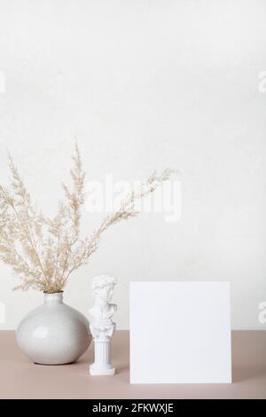 Carta bianca, mockup di invito con piccola statua e pampas erba in vaso su sfondo bianco e beige. Arredamento moderno. Spazio copia Foto Stock