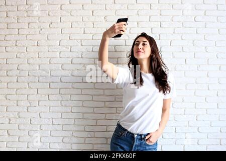 Bella giovane donna, lunghi capelli castani ondulati, vestita in stile anni '90, facendo selfie. Donna che indossa jeans mom e maglietta bianca normale che scatta le foto di Foto Stock