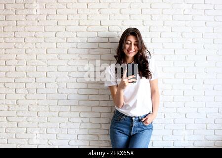 Giovane donna attraente che indossa una t-shirt bianca vuota che riceve un messaggio sul nuovo cellulare. Bel modello femminile, sorridente, tenendo in mano il gadget mobile, Foto Stock