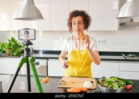 La donna youtuber si registra con il suo smartphone in cucina mentre si prepara una ricetta di insalata, c'è luce naturale e lei indossa una a gialla Foto Stock