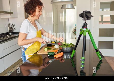 La donna youtuber si registra con una macchina fotografica nella cucina della sua casa mentre prepara una ricetta di insalata, c'è luce naturale e lei indossa un giallo Foto Stock