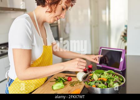 Giovane donna caucasica con capelli ricci prepara un'insalata nella cucina della sua casa mentre guarda il suo tablet digitale, è luce del giorno e c'è na Foto Stock