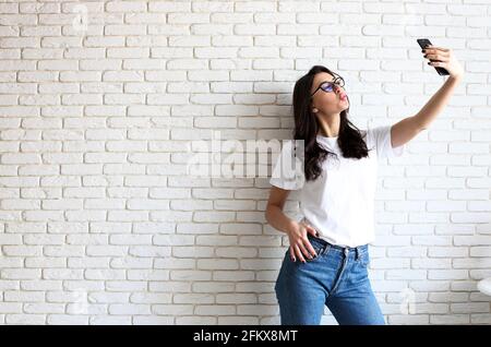 Bella giovane donna in occhiali da vista gatto, facendo selfie su dispositivo mobile gadget. Donna Hipster che indossa jeans mom e camicia bianca normale che scatta le foto Foto Stock