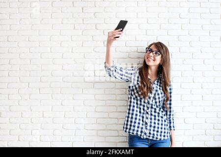 Bella giovane donna, lunghi capelli castani ondulati, vestita in stile anni '90, facendo selfie. Donna che indossa jeans mamma e camicia a scacchi scattando foto di lei Foto Stock