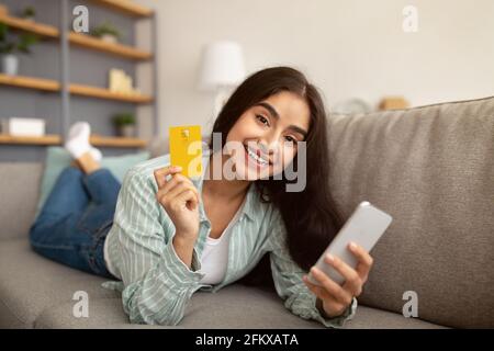 Shopping online. Signora indiana con smartphone e carta di credito sdraiato sul divano a casa, l'acquisto di merci nel negozio web Foto Stock