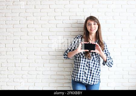 Giovane attraente donna che mostra il suo nuovo touch cellulare. Concentratevi sulla mano e sullo smartphone. Bella modello femminile su sfondo bianco mattone Foto Stock