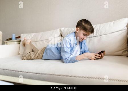 Il bambino giace sul divano nelle mani che tiene un telefono, gioca con esso. Foto Stock