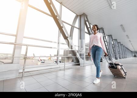 Happy African American Traveller Woman Walking con valigia in Terminal dell'aeroporto Foto Stock