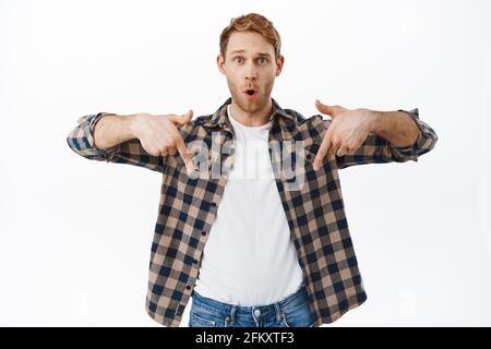 Uomo attraente sorpreso con i capelli rossi, gastping dire WOW, puntando le dita verso il basso a incredibile nuova offerta promozionale, miglior affare in negozio, mostrando pubblicità Foto Stock