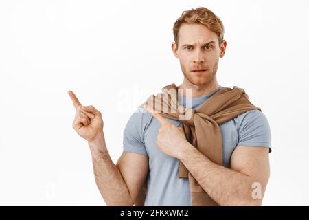 Uomo rosso sospetto e scettico che ha dubbi e fa domande su un elemento, puntando all'angolo in alto a sinistra qualcosa di strano, accigliato dubbio Foto Stock