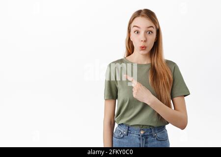 Cute autentica ragazza guarda sorpreso e intrigato mentre i punti a sinistra, mostra pubblicità promozionale testo da parte e fissa a macchina fotografica curioso, si alza Foto Stock