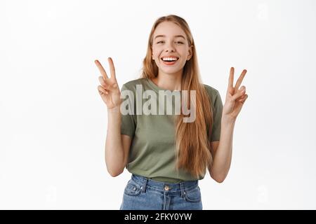 Immagine di bella ragazza bionda, sorridente e sorridente felice, mostrando pace segno v, kawaii in piedi su sfondo bianco Foto Stock