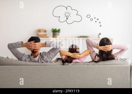 Giovane famiglia di tre seduti insieme sul divano con le braccia dietro la testa e la donna sognare, avendo nuove idee Foto Stock