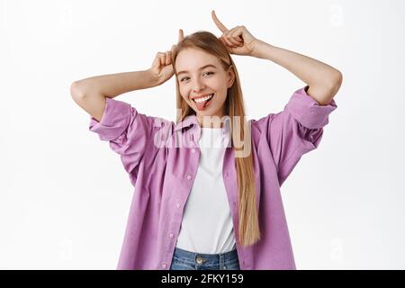 Divertente ragazza bionda, mostra corna toro diavolo gesto, mostrando lingua e sorridente sciocco, in piedi su sfondo bianco Foto Stock