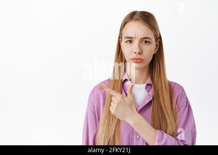 Primo piano di triste e deluso ragazza bionda che punta il dito a sinistra, sulking e lamentando, mostrando qualcosa di cattivo ingiusto, in piedi su bianco Foto Stock