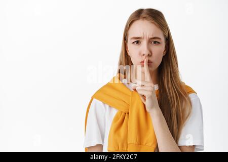 Primo piano di bionda giovane donna che gridano, premere il dito sulle labbra e guardare sconvolto, hushing, dire shhh dicendo di mantenere il silenzio, in piedi contro bianco Foto Stock