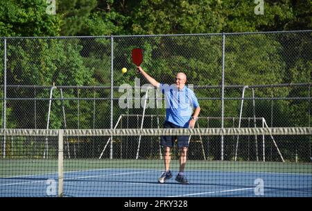 Il maschio anziano restituisce un hit alto mentre gioca a Pickle Ball, su un campo da tennis esterno. Ha su una camicia a righe blu. Foto Stock