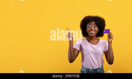 Concetto di pubblicità. Donna afroamericana che tiene la carta di credito e che punta allo spazio vuoto su sfondo giallo Foto Stock