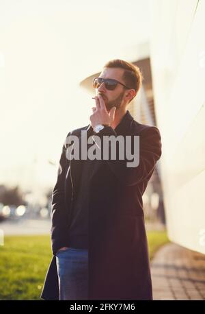 L'uomo elegante in un cappotto e bicchieri fuma una sigaretta Foto Stock