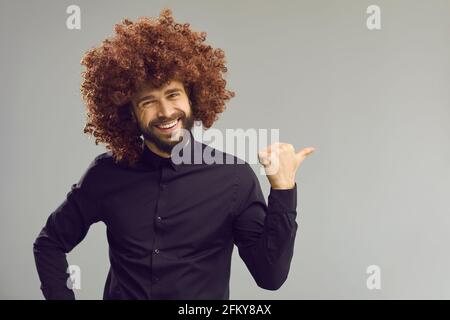 Uomo divertente in parrucca arricciata che punta il pollice da parte a vuoto sfondo dello spazio di copia pubblicitario Foto Stock