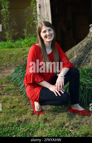 La giovane donna si inginocchia nel suo cortile e sorride. Si trova di fronte ad un albero e ad un granaio rustico di legno. Indossa una camicia rossa e dei jeans neri. Foto Stock