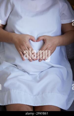 Giovane donna asiatica incinta che fa un cuore con le mani sul ventre Foto Stock