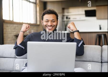 Felice eccitato ragazzo indiano che celebra la vittoria, guarda il portatile seduto sul divano a casa, leggendo buone notizie, fortunato sopravorato uomo di corsa mista che solleva pugni in gesto trionfale Foto Stock