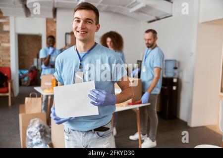 Ritratto di giovane volontario maschio caucasico in uniforme blu e guanti protettivi sorridenti alla macchina fotografica, tenendo in bianco banner di carta per il tuo annuncio Foto Stock