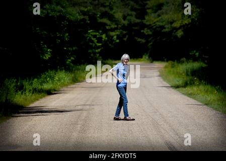Una donna di novantanove anni cammina lungo una strada di campagna deserta. Forest si chiude su di lei, ma il suo spirito indomabile rifiuta di cedere. Mani sui fianchi Foto Stock