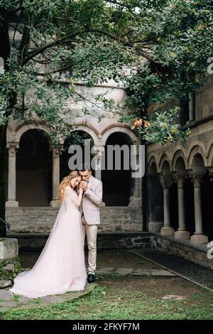 Lo sposo bacia le mani della sposa mentre si trova in un giardino verde sullo sfondo di un'antica villa con colonne sul Lago di Como Foto Stock