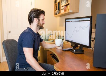 Uomo che guarda la tabella di finanza sul calcolatore, commerciante di giorno, che investe dalla sede, sorseggiando il caffè, uomo disteso Foto Stock