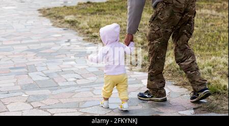 La bambina va mano in mano con il papà all'aperto. L'uomo militare in tute conduce il bambino a mano. Giorno della memoria. Festa del Padre. Buona famiglia. Por Foto Stock