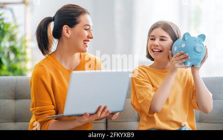 Madre e figlia tengono una banca piggy e un computer portatile seduti sul divano a casa. Foto Stock