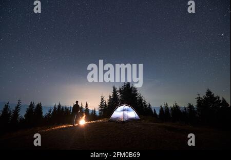 Vista del cielo stellato notturno sulla valle di montagna con escursionista maschile, tenda illuminata campeggio e falò. Uomo viaggiatore in piedi vicino tenda turistica sotto il cielo notturno con stelle. Concetto di campeggio notturno. Foto Stock