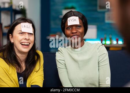 Amici multirazziali che si godono l'amicizia mentre si gioca a indovinare chi gioco attaccando note appiccicose sulla fronte. La gente di corsa mista che si diverte, ridendo insieme mentre si siede sul divano a tarda notte. Foto Stock