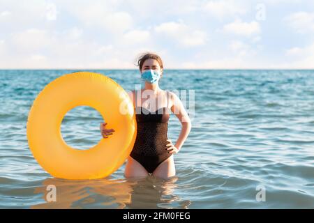 Distanza sociale e protezione da virus. Una donna in costume da bagno e maschera medica si pone con un cerchio gonfiabile in piedi in mare. Nel backgroun Foto Stock