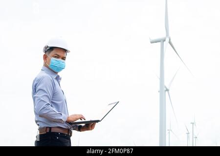 Ingegnere mulini a vento indossando maschera e lavorando su computer portatile con la turbina eolica in background Foto Stock